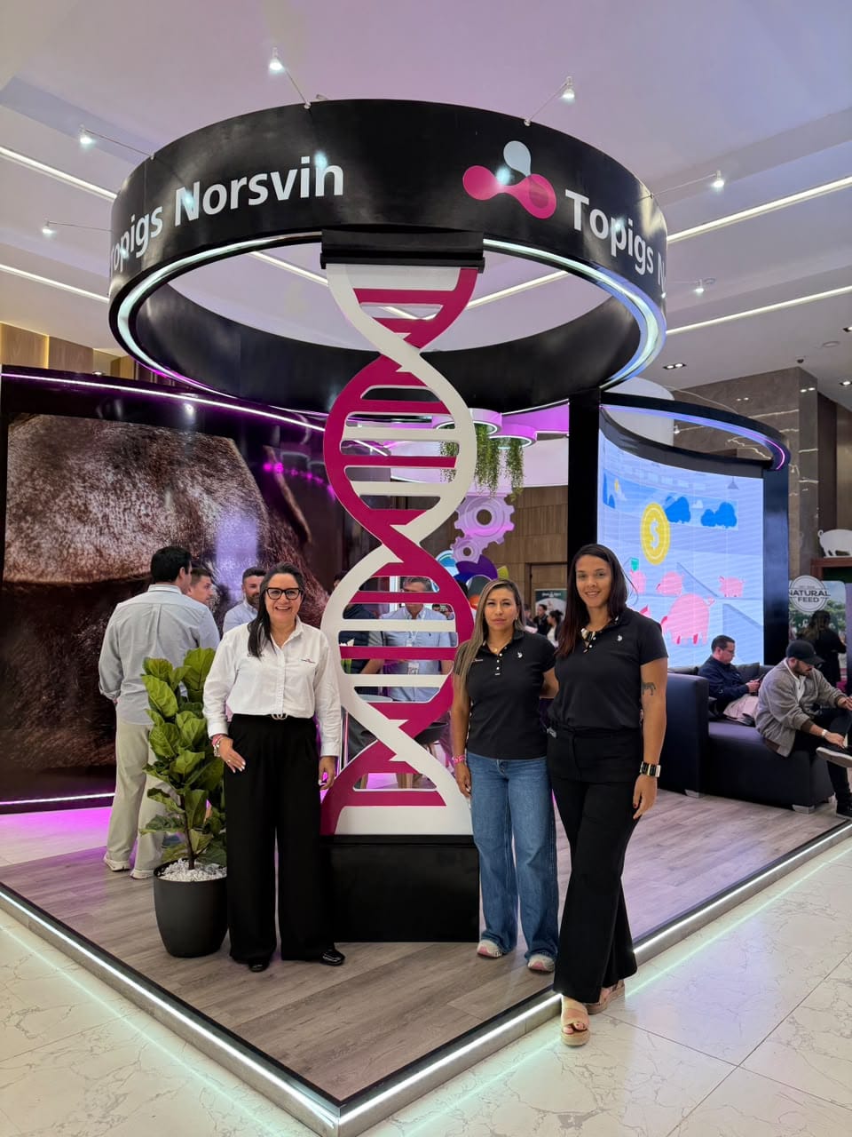 Tres personas posan delante del stand de Topigs Norsvin, junto a una gran hélice de ADN y bajo un anillo con el logotipo de la marca
